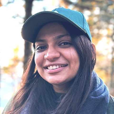 Dhanshri smiling in a green cap and blue scarf, with autumn trees behind her.