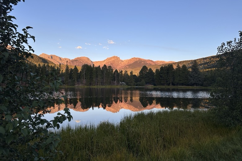 Mountain peaks glowing orange at sunset, mirrored in Sprague Lake, edged with pines and tall grass.