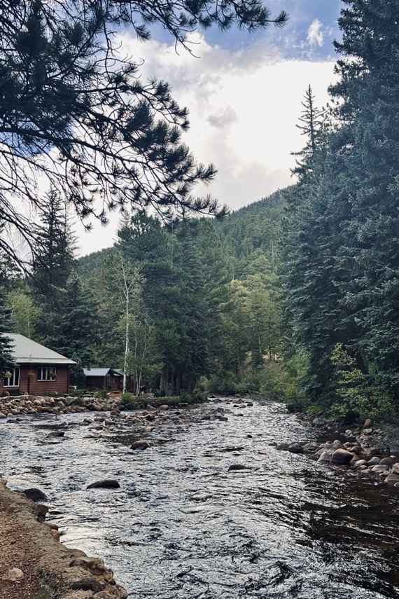 A rocky mountain river running past a wooden cabin, with pine trees on both banks, in Estes Park.