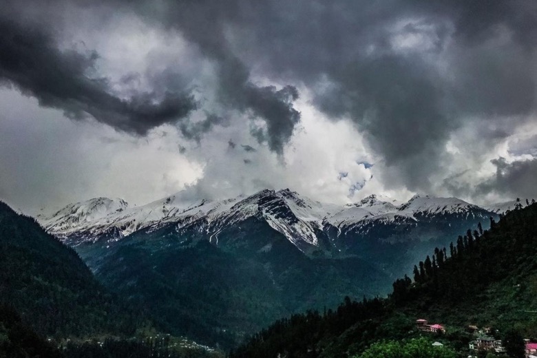 Snow-covered peaks under dark storm clouds, above a forested valley with a few houses, in Tosh.